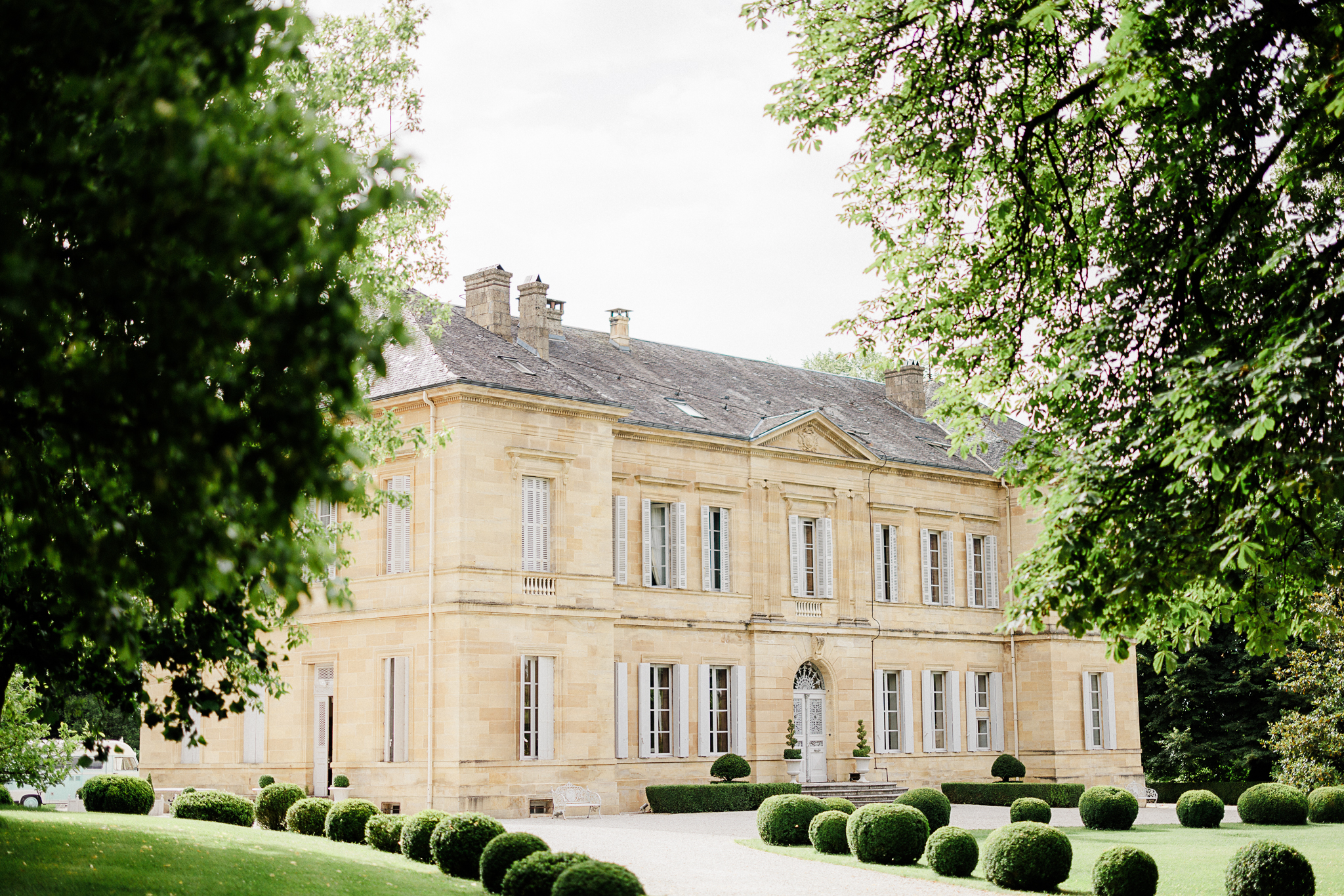 A beautiful chateau with manicured gardens and trees framing the entrance. A vintage camper van is parked on the left side of the image, suggesting a possible event or gathering.