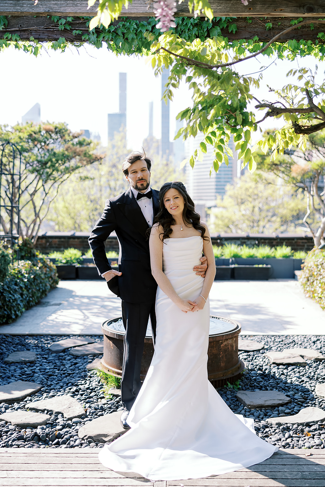 A bride in a strapless white wedding gown and a groom in a black tuxedo stand together on a lush rooftop garden terrace with city skyscrapers in the background.