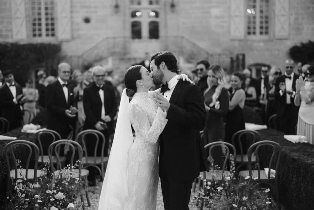 A bride in a lace dress and a groom in a tuxedo kiss during their wedding ceremony. Guests are seated in the background.