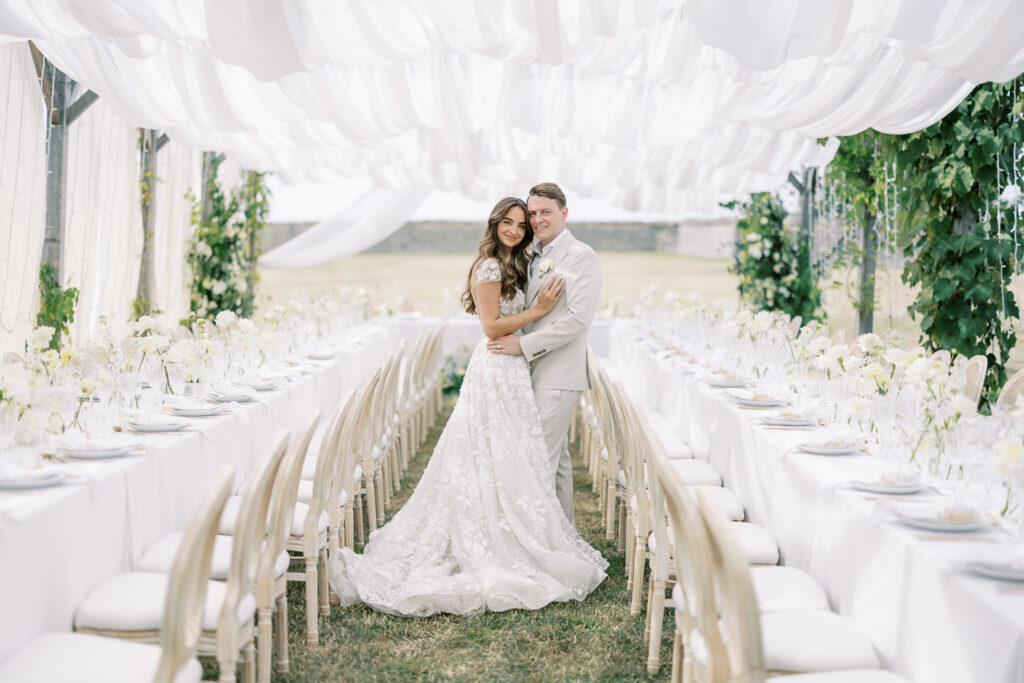 A bride and groom embrace under a draped canopy at an outdoor wedding reception. Long tables are set with white linens, plates, and floral arrangements.